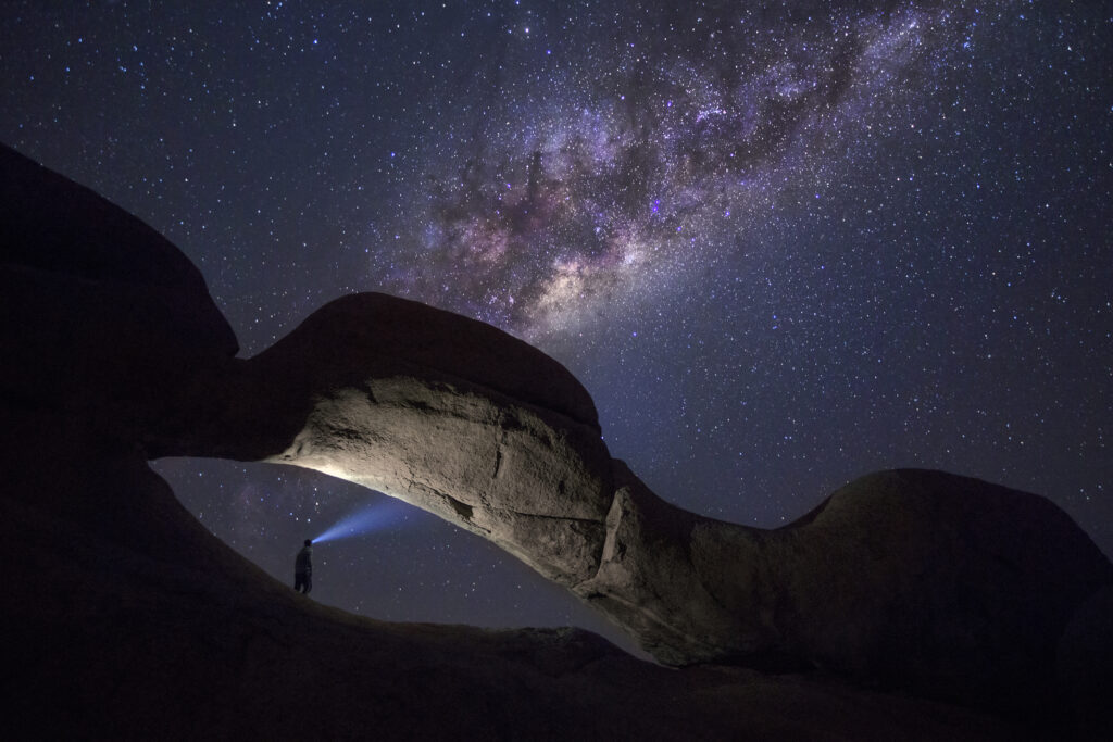 A person stands staring at the milky way from the rock arch at Spitzkoppe.